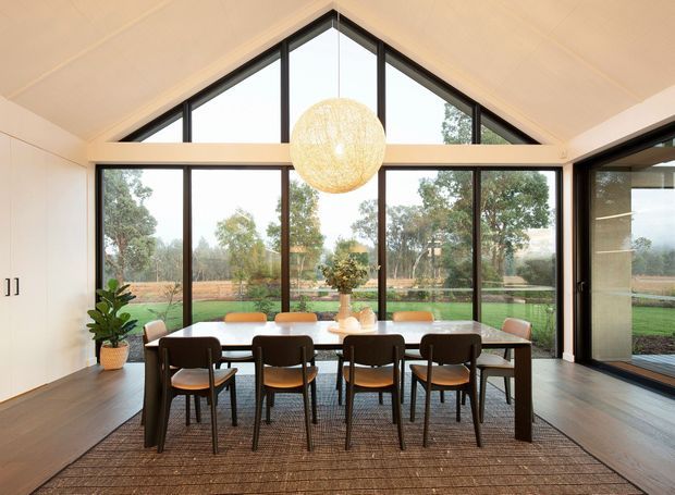 A bright dining room with a long dark timber dining table and matching chairs, positioned in front of expansive black-framed glazing. The room has a high pitched ceiling with a triangular clerestory window, warm timber flooring, white walls, and a large woven spherical pendant light. A landscaped rural garden is visible through the windows.