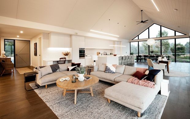 Open-plan living, dining and kitchen area with a large sectional sofa and ottoman in the foreground, a round timber coffee table, textured area rug, and a bright white kitchen with island seating behind. Full-height black-framed glazing and a dramatic gabled glazed wall bring in abundant natural light. The room has warm timber flooring, white cabinetry and walls, exposed sloped ceiling planes, and a contemporary relaxed residential design.