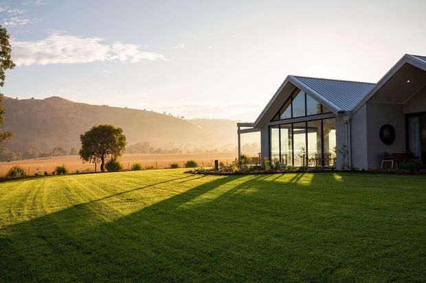 A contemporary rural house set beside a broad, freshly mown lawn, with dark pitched metal roofs, pale rendered walls, and a prominent glazed gable pavilion. The home overlooks open golden fields and wooded hills in warm low-angle sunlight, creating a spacious countryside setting.