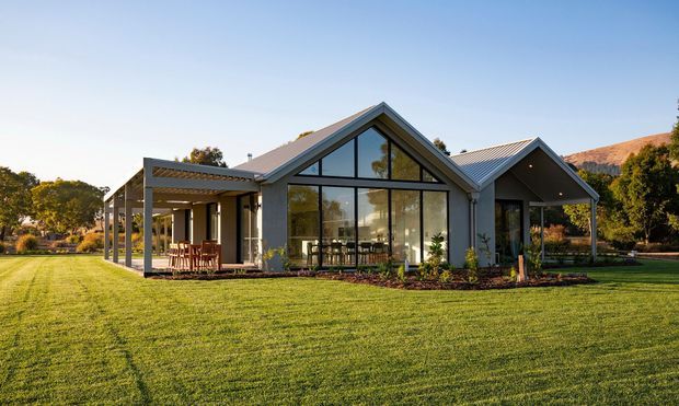 A contemporary single-storey home set within a broad landscaped lawn, featuring interconnected pitched gable roofs, extensive floor-to-ceiling glazing, and a covered outdoor dining terrace. The facade uses dark grey cladding and framing with warm timber details, while the transparent central gable reveals the living and dining areas. The property is surrounded by mature trees and rolling hills in the background.