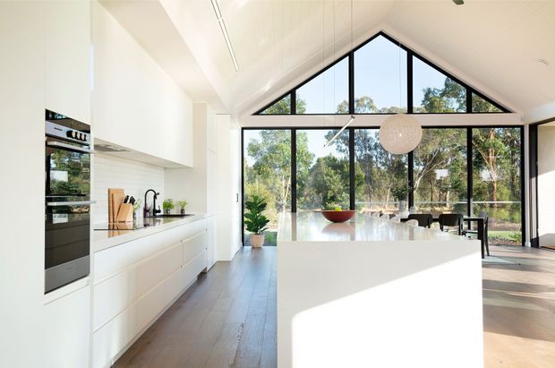 Bright open-plan kitchen and dining area with a large white island, streamlined white cabinetry, integrated stainless-steel appliances, and a timber floor. Dramatic triangular clerestory glazing and full-height windows bring in abundant daylight and overlook leafy trees. A spherical pendant light hangs above the dining table, reinforcing the clean contemporary feel.