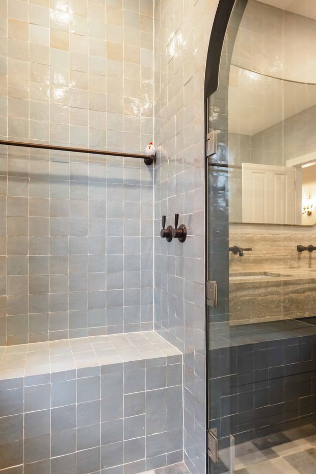 A close view of a tiled bathroom shower enclosure with pale blue-grey and cream square wall tiles, a built-in tiled bench, dark bronze shower controls and a dark-framed glass shower door. A stone vanity with dark tiled cabinetry is visible through the glass and mirror reflection.