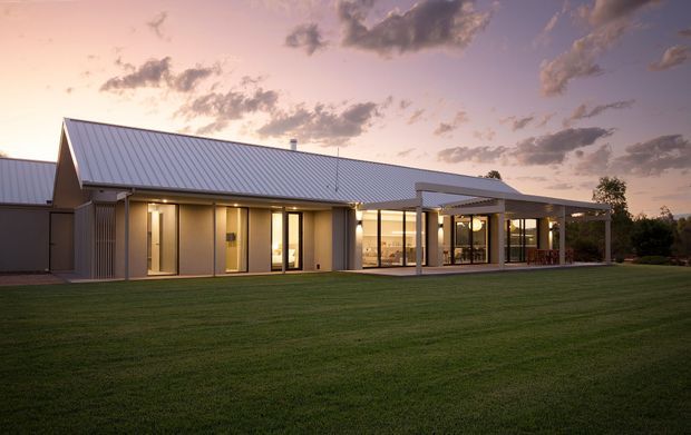 A single-storey contemporary house photographed at dusk, with a long low-profile form, prominent pitched metal roof, pale vertical-panel exterior walls, extensive floor-to-ceiling glazing, and a covered outdoor terrace extending from the living areas. The building sits behind a broad manicured lawn, with warm interior lighting contrasting against the purple evening sky.