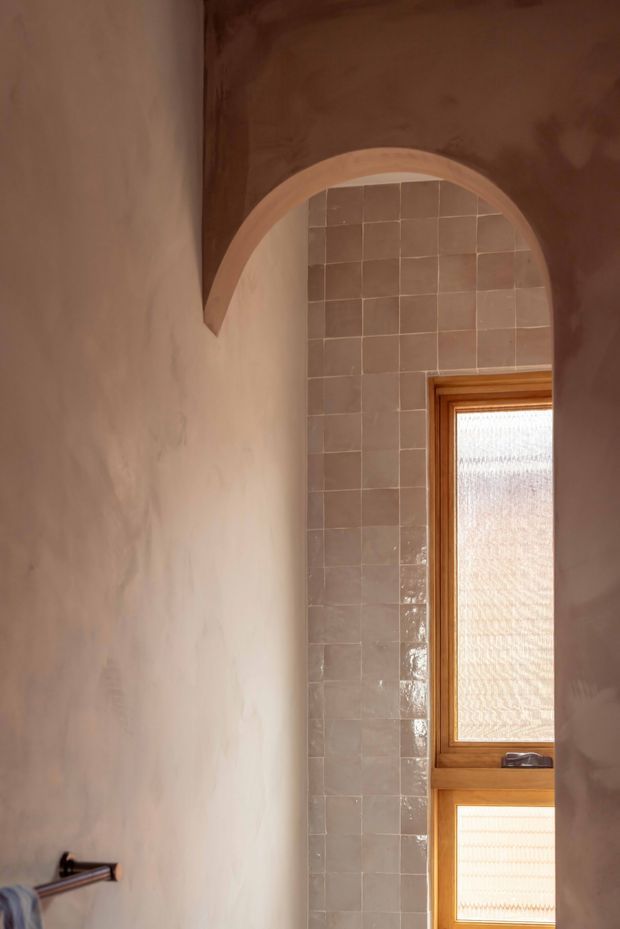 A warm, intimate interior detail featuring a softly plastered wall and an arched opening framed with timber. The opening reveals a vertical tiled wall in muted taupe-grey square tiles and a timber-framed textured-glass window. A small metal wall fixture is visible in the lower left. The image emphasizes handcrafted surfaces, curved architectural detailing, warm timber, and subdued natural light.