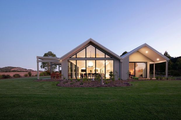 Single-storey gabled rural-style residence photographed at dusk, with a central glazed living space, an attached gabled wing, and a covered outdoor area. The home has light-coloured rendered or painted walls, dark pitched roof edges, extensive floor-to-ceiling glazing, warm interior lighting, and a broad manicured lawn with landscaped garden beds.