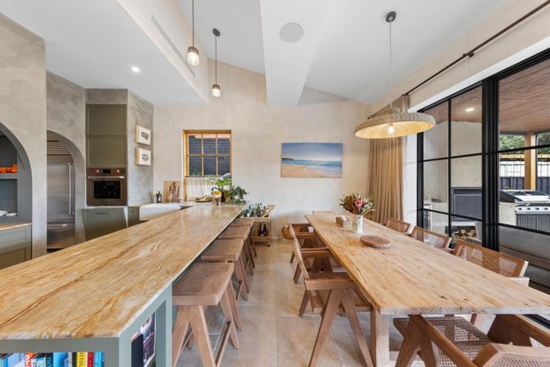 Bright open-plan kitchen and dining area with a large natural stone island, fixed cabinetry, timber dining table and benches, warm woven pendant lighting, arched wall openings, and black-framed glazing leading to an outdoor barbecue area. The space has a warm, relaxed contemporary coastal feel.