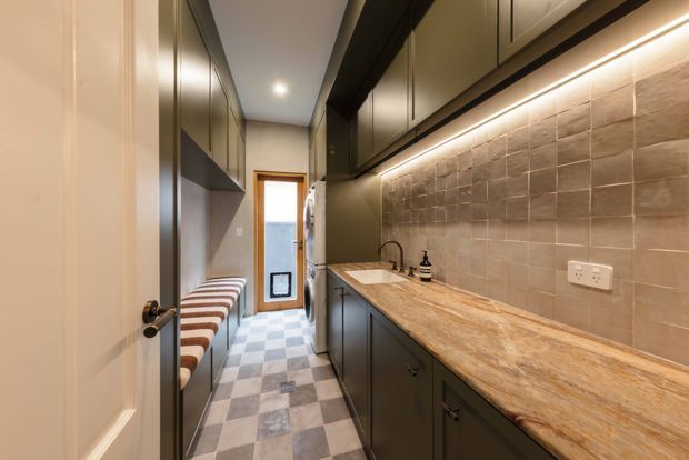 A narrow, well-organised laundry or utility room with dark built-in cabinetry, a long timber-look benchtop, integrated sink, warm under-cabinet lighting, and a textured neutral tiled splashback. The opposite wall includes built-in seating or storage, while the checkered tile floor leads toward a glazed exterior door.
