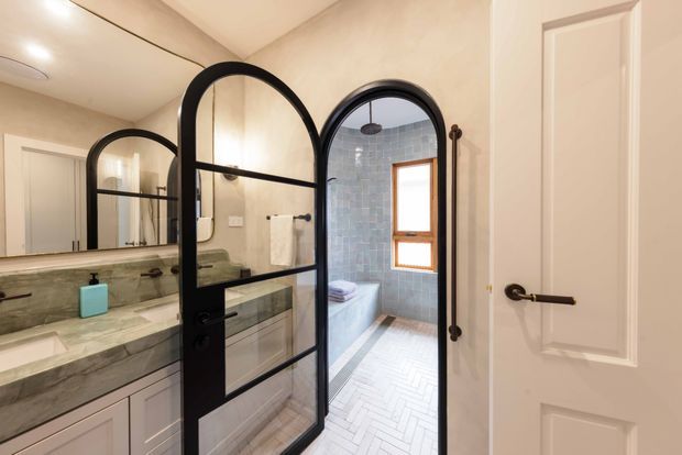 A refined bathroom interior viewed from the entry, featuring a double vanity with pale marble-look benchtop and integrated basins, a large wall mirror, and distinctive black-framed arched glass doors. The doors open to a tiled shower or bathing area with blue-grey wall tiles, a timber-framed window, and a light herringbone floor. Warm neutral walls, white cabinetry, dark hardware, and arched detailing create a classic yet contemporary feel.