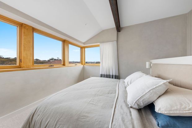 Bright, minimalist bedroom with a low-profile bed dressed in layered white and pale grey bedding. Warm timber-framed corner windows provide expansive views, while a dark exposed ceiling beam, pale plaster walls, and sheer curtains add understated architectural character.
