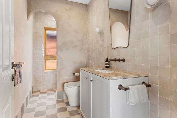 A compact, warmly styled bathroom with a toilet, single vanity, tiled walls, checkerboard floor tiles, arched openings, and a small timber-framed window. The vanity has pale cabinetry, a timber-toned benchtop, and dark metal fixtures, while a wall-mounted curved mirror and exposed plumbing details add character.