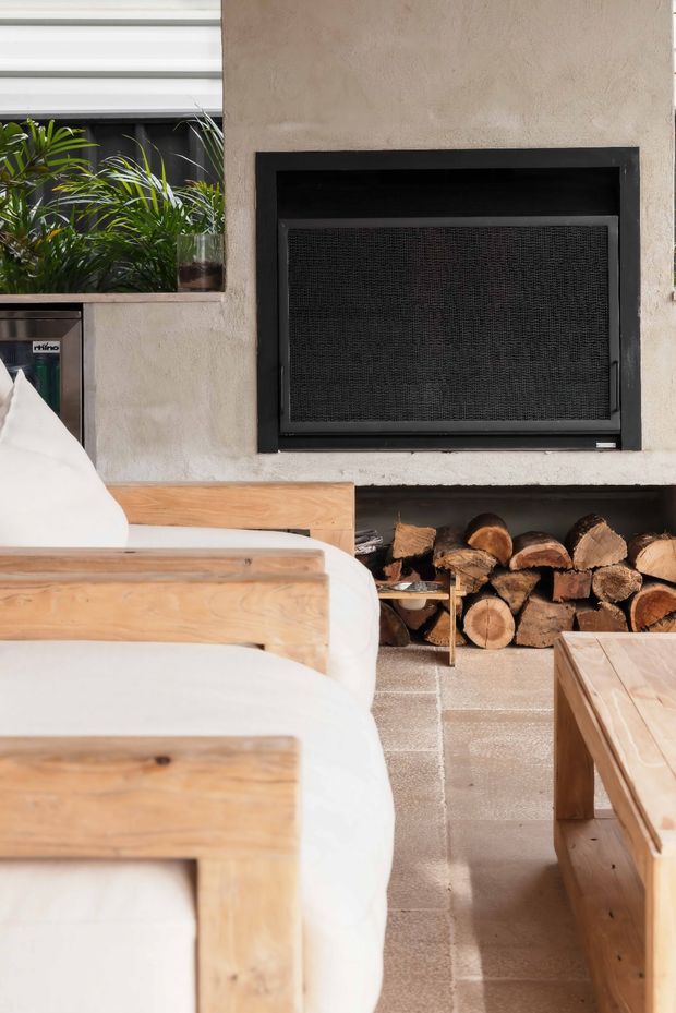 A warm, contemporary living room focused on a built-in fireplace with a black metal firebox and open timber log storage beneath. Pale textured plaster walls, a large horizontal window with dark framing, natural timber-framed white upholstered seating, a timber coffee table, and large-format neutral floor tiles create a relaxed, earthy interior.