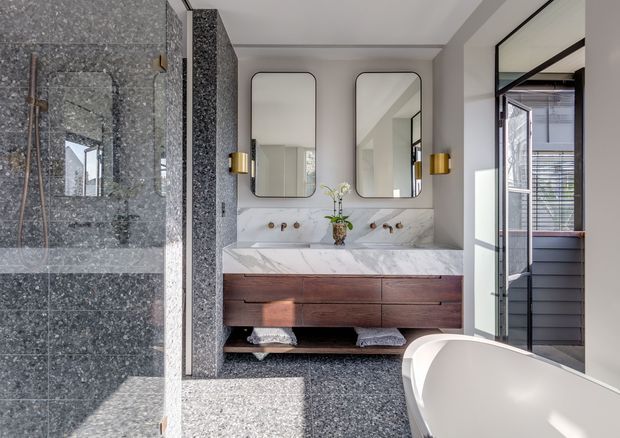 A refined contemporary bathroom with a double vanity, freestanding bath, and walk-in shower. The room features dark speckled stone or terrazzo-like wall and floor surfaces, a white veined stone vanity benchtop and splashback, warm timber vanity drawers, rounded rectangular mirrors, brass wall sconces and tapware, and black-framed glazing opening toward an exterior area. Natural light creates a bright yet sophisticated atmosphere.