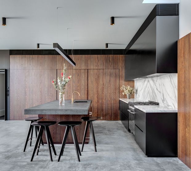 A sophisticated contemporary kitchen with a large grey stone island and black bar stools, surrounded by dark timber wall cabinetry and panelling. The kitchen includes a black appliance wall, a veined white marble-look splashback, dark cabinetry, integrated storage, and minimalist black pendant and recessed lighting. The polished concrete floor and warm timber create a refined, high-contrast interior.
