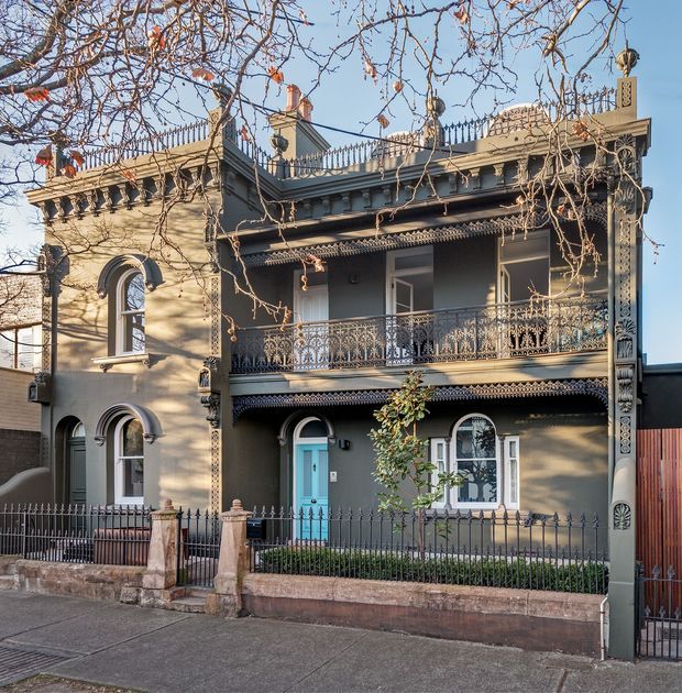 Exterior street view of a two-storey Victorian-era terrace or townhouse with an ornate rendered facade, decorative cast-iron balconies and roofline cresting, arched windows, elaborate column details, and a small fenced front garden. The facade is painted in muted grey and beige tones with a contrasting turquoise front door.
