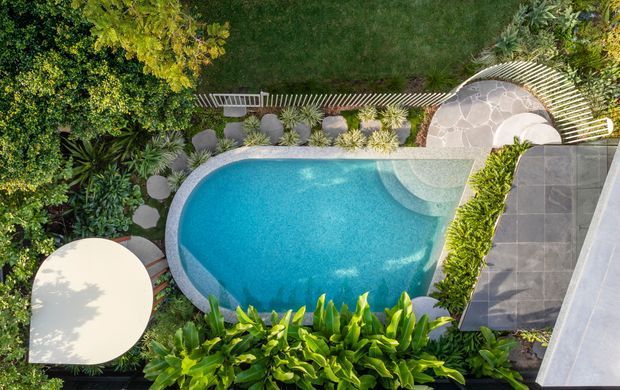 Aerial view of a landscaped residential pool area. A curving turquoise swimming pool is surrounded by lush tropical planting, rounded stepping stones, a pale paved terrace, dark grey tiled paving, and a slatted privacy screen. A rounded outdoor dining table is visible at the lower left.