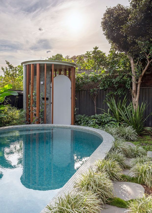 A landscaped residential pool area with a curved turquoise swimming pool edged in small grey tiles. Beside the pool is a compact curved outdoor pool pavilion or changing-room structure with vertical timber slats, translucent panels, and a distinctive rounded white door beneath a shallow curved roof. Dense planting, ornamental grasses, mature trees, and dark fencing create a private garden setting.