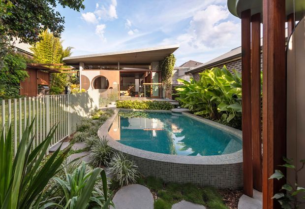A lush, landscaped courtyard with a curved tiled swimming pool as the focal point. The pool is enclosed by a slender metal fence and surrounded by dense tropical planting, stepping stones and garden beds. Beyond it is a compact modern pavilion or outdoor living structure with timber-framed glazing, circular wall openings and a broad low-pitched roof. Warm timber elements contrast with pale rendered surfaces and abundant greenery.