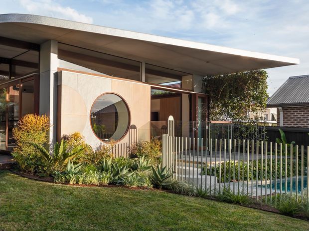 Single-storey modern residential facade with a broad low-slung flat roof, extensive glazing, and a distinctive pale timber or timber-look entry screen featuring a large circular window. A landscaped front garden with grasses and sculptural planting sits behind a slim vertical metal fence, with a paved path leading to the entrance. A neighbouring brick house is visible in the background.