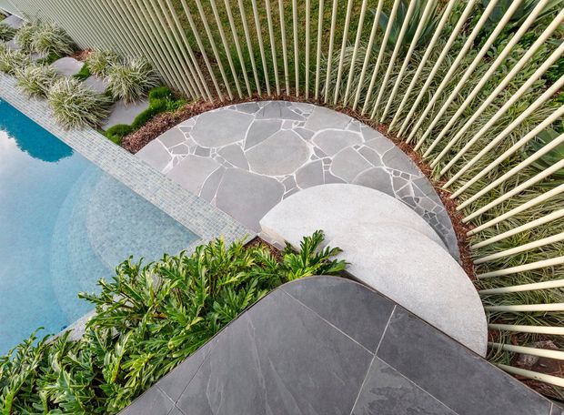 Overhead view of a landscaped pool area with curved organic forms. A pale blue swimming pool borders an irregular natural-stone paved terrace and a rounded light concrete platform, with dark charcoal paving in the foreground. Dense tropical planting and a tall curved vertical timber or bamboo screen enclose the space.