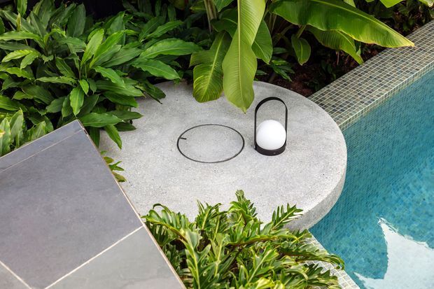 Close-up view of a landscaped poolside setting featuring a curved light-grey concrete platform beside a tiled swimming pool. Tropical foliage surrounds the space, with a small black-and-white portable light on the platform and large-format grey paving or tiles in the foreground.