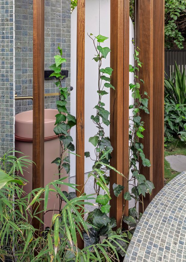 A lush outdoor bathroom or poolside bathing area viewed through vertical timber slats and climbing greenery. The space features small grey mosaic wall tiles, a partially visible freestanding pink-toned bathtub, dark tapware, white rendered walls, and a curved grey mosaic-tiled surface in the foreground. Dense planting surrounds the timber privacy screen, creating a secluded garden setting.