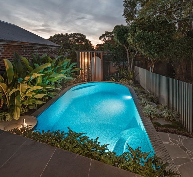 A landscaped residential courtyard at dusk featuring a compact, curved swimming pool with vivid turquoise water, surrounded by lush tropical planting, dark paving, a corrugated metal privacy fence, and a brick building with a dark pitched roof. A small illuminated pool entry or outdoor shower structure stands at the far end, creating a warm contrast against the cool pool lighting.