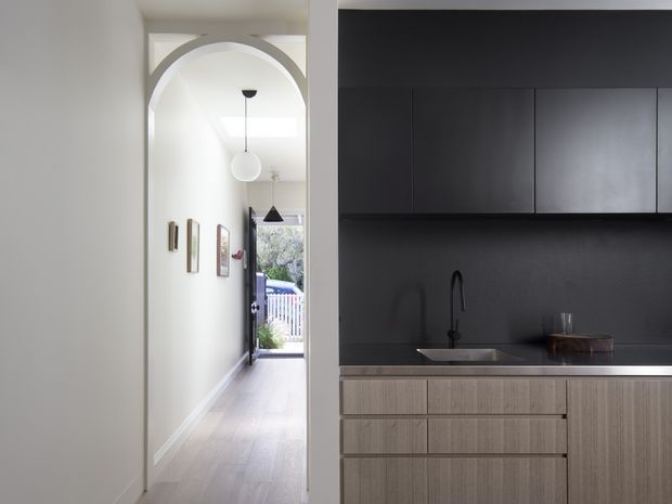 A compact contemporary kitchen viewed beside a long hallway. The kitchen features dark matte upper cabinetry and a matching dark splashback, light wood-grain lower cabinetry, a slim stone benchtop, and a black undermount sink with a tall tap. An arched opening leads into a bright hallway with pale flooring, white walls, framed artwork, pendant lights, and an exterior doorway at the far end.