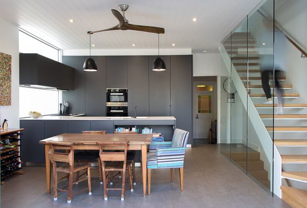 Open-plan contemporary kitchen and dining area with dark full-height cabinetry, a pale island bench, integrated appliances, timber dining table and chairs, and a colourful striped armchair. A glazed staircase with floating timber treads rises along the right side, beneath a white panelled ceiling with recessed lighting and a sculptural ceiling fan.