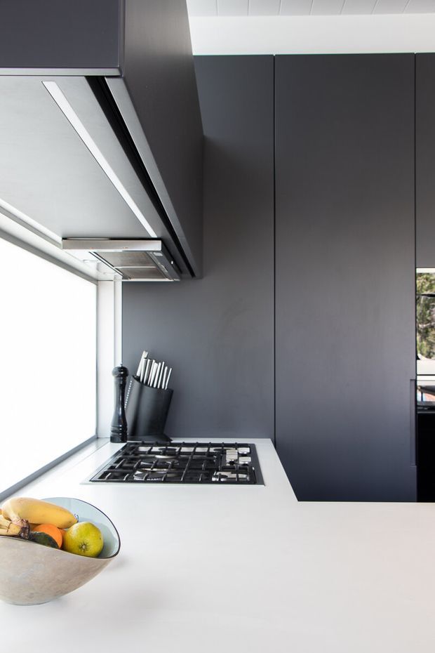 A contemporary kitchen featuring a white island benchtop with an integrated gas cooktop, tall matte dark-grey cabinetry, and a stainless-steel rangehood. A large window provides bright natural light, while a fruit bowl and knife block sit on the island. The palette is clean and strongly contrasted between white surfaces and charcoal cabinetry.