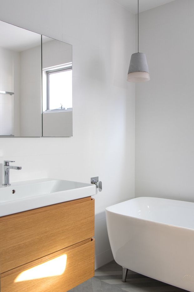 Bright contemporary bathroom with a freestanding white bathtub, wall-mounted white basin, chrome tapware, mirrored wall cabinet, pale tiled walls, grey floor tiles, and warm timber vanity cabinetry. A simple grey pendant light hangs above the bath.