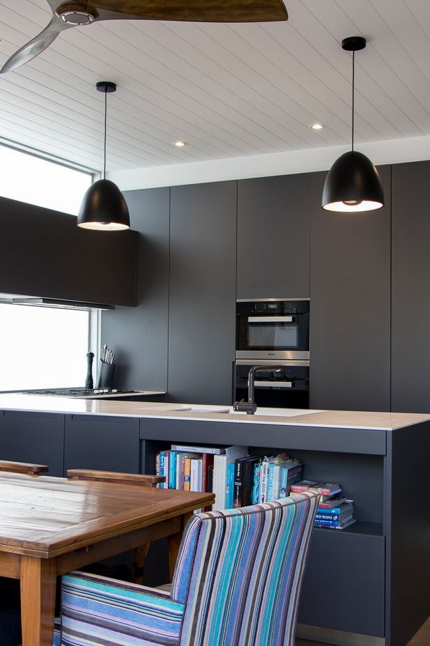 A contemporary open-plan kitchen and dining area with full-height matte dark cabinetry, integrated stainless-steel ovens, a light benchtop, and open shelving filled with books. A timber dining table and striped upholstered chair occupy the foreground. The space has a white-lined ceiling, black pendant lights, a ceiling fan, and large windows providing abundant natural light.