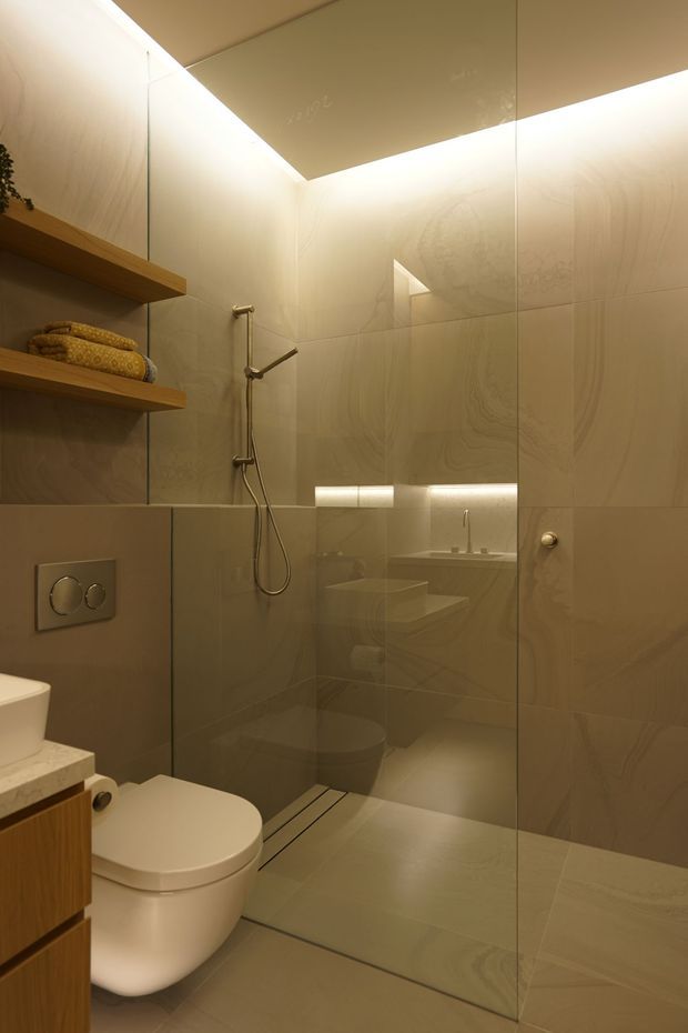 A compact, refined bathroom with a walk-in glass shower, wall-mounted toilet, floating timber vanity, open timber shelving, and concealed warm LED lighting. Large-format beige and taupe stone-look wall and floor tiles create a calm, continuous finish, complemented by brushed metal shower fittings and understated sanitaryware.