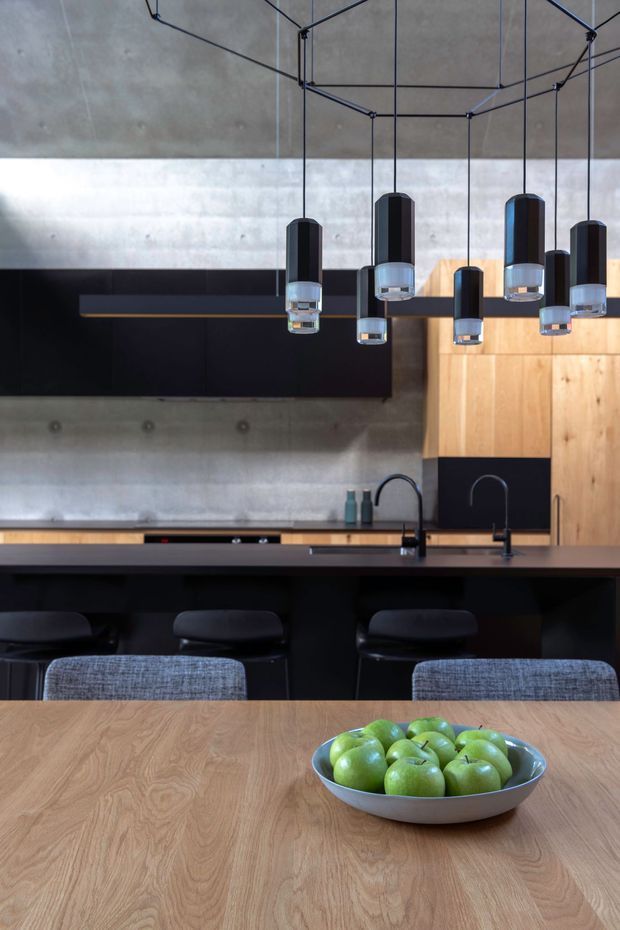 A contemporary open-plan kitchen and dining area with a dark island bench, black bar stools, timber cabinetry, concrete walls, and a large black geometric pendant light installation. A light timber dining table with upholstered chairs and a bowl of green apples occupies the foreground.