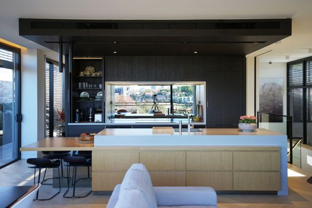 Contemporary open-plan kitchen viewed from the adjoining living area, featuring a large pale timber island with a light benchtop, dark timber cabinetry and ceiling treatment, integrated shelving, bar stools, and broad glazed openings with blinds. A dining and living area are visible in the foreground, with an urban outlook beyond the kitchen.