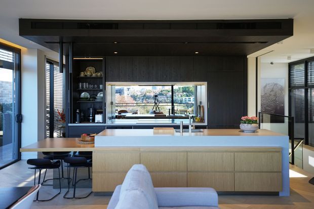 A contemporary open-plan kitchen with a large central island, integrated light timber cabinetry, dark timber wall and ceiling joinery, and a framed view through the rear glazing. Black bar stools sit at the island, with extensive windows and daylight creating a bright, refined interior.
