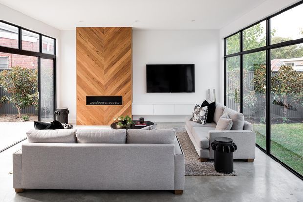 Bright contemporary living room with floor-to-ceiling black-framed glazing on two sides, a white minimalist interior, polished concrete flooring, and a prominent herringbone timber feature wall incorporating a linear fireplace. Two light-grey sofas face a wall-mounted television, with a neutral rug and restrained black accents.