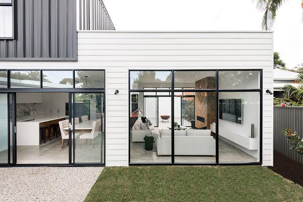 Exterior view of a contemporary two-storey residential extension with white horizontal weatherboard cladding, dark vertical metal cladding above, and extensive black-framed floor-to-ceiling glazing. The glazing reveals a bright open-plan kitchen and living room, while a lawn, gravel path, fencing, and tropical planting frame the facade.