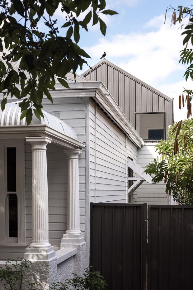 Exterior view of a compact, heritage-influenced residential building framed by mature foliage. The facade combines light grey horizontal weatherboards with a steep gabled upper volume clad in vertical boards, detailed classical fluted columns and moulded trim at the entry, and a curved corrugated metal porch roof. A dark privacy fence occupies the foreground, while a small upper-level window and roofline are visible behind.