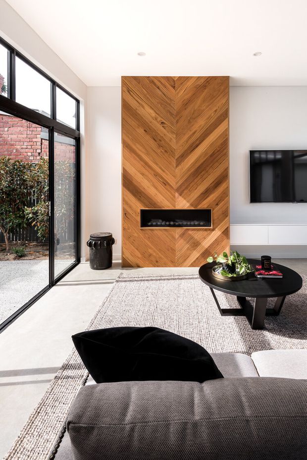 Bright contemporary living room with a large black-framed glazed opening, pale walls and floor, and a prominent floor-to-ceiling chevron-patterned timber feature wall incorporating a built-in fireplace. A wall-mounted television, low white joinery, round black coffee table, textured rug and dark sofa complete the space. The glazing opens toward a small landscaped courtyard and exposed brick wall.