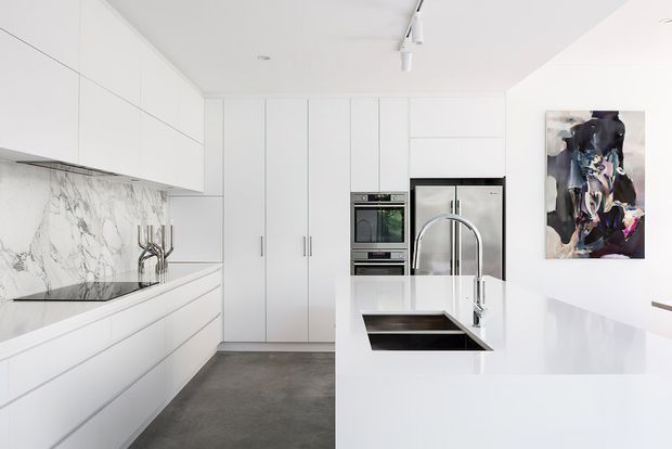 Bright, minimalist contemporary kitchen with extensive white flat-panel cabinetry, integrated appliances, a large white island with a dark inset sink and curved chrome tap, and a dramatic white marble-look splashback. The room has a polished, uncluttered appearance with grey flooring and a colourful abstract artwork on the wall.