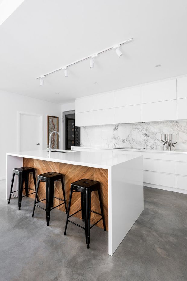 Bright contemporary kitchen featuring a large white island with waterfall ends, integrated sink and tap, timber-look panelled breakfast bar, and three black metal stools. The room has extensive white handleless cabinetry, a dramatic white-and-grey marble-look splashback, polished concrete flooring, recessed lighting, and a white track light.