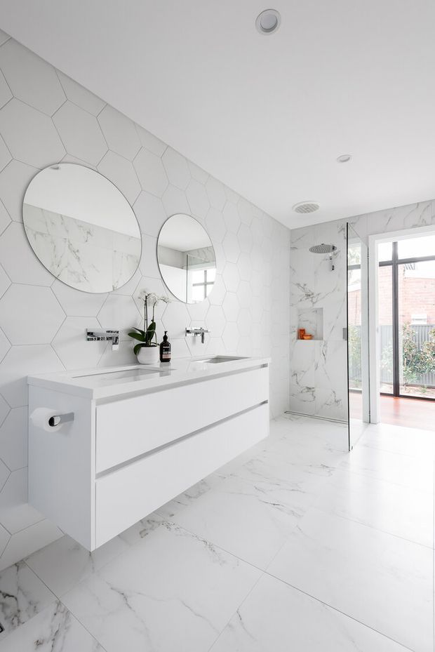 Bright contemporary bathroom with a wall-mounted double vanity, white handleless drawers, twin round mirrors, and a walk-in shower. The walls feature white geometric hexagonal tiles, while the shower enclosure and floor use large white marble-look tiles with grey veining. A glazed doorway opens to an exterior deck or patio area.