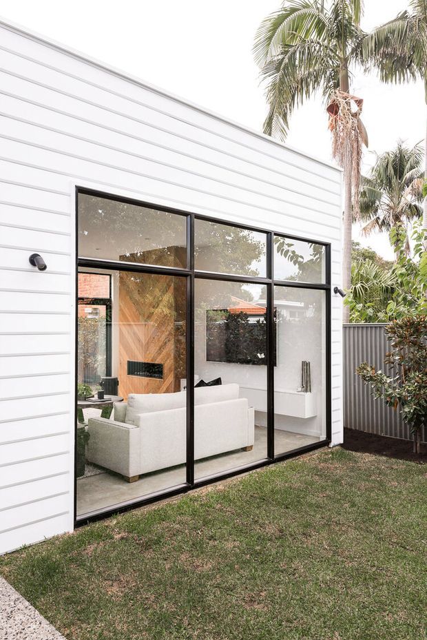 Exterior view of a contemporary home extension with white horizontal cladding and a large black-framed glazed wall. Through the glazing, a bright living room is visible with a pale sofa, timber feature wall, fireplace, and white built-in media cabinetry. A lawn, palm trees, and a corrugated metal boundary fence surround the exterior.
