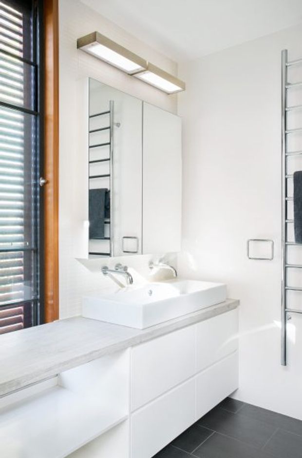 Bright, compact contemporary bathroom featuring a white floating vanity with integrated drawers, a square countertop basin, wall-mounted tapware, large mirrored cabinet, and a horizontal wall light. Dark charcoal floor tiles contrast with the predominantly white surfaces. A heated towel rail and a timber-framed louvred window are visible.