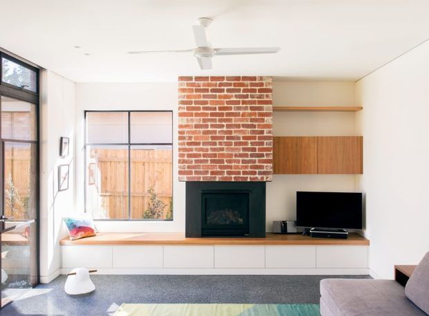 Bright contemporary living room with a built-in media and fireplace wall. A red exposed-brick chimney breast is centered above a dark fireplace, flanked by white built-in cabinetry, open shelving, and warm timber joinery. Full-height glazing and a large window bring in abundant daylight, while a ceiling fan, grey carpet, low seating, and a television complete the room.