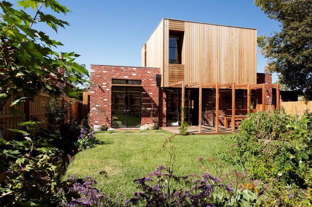 A contemporary two-storey residential extension viewed from a landscaped garden. The lower level features exposed red brick walls, dark-framed glazing and a covered outdoor area, while the upper level is a projecting timber-clad volume with vertical boards and a tall narrow window. The garden includes lawn, mature planting and flower beds under clear daylight.