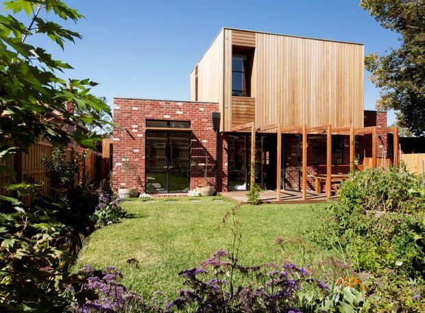 A contemporary two-storey residential extension viewed from a landscaped garden. The lower level combines exposed red brick walls with extensive dark-framed glazing and a timber-framed covered deck or verandah, while the upper level is a simple box clad in vertical light timber boards. The garden includes lawn, planted borders, mature vegetation and flowering plants under a clear blue sky.