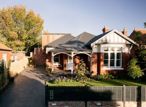 Street-facing view of a single-storey heritage-style brick house with a steep gabled roof, prominent front gable, decorative white timber veranda framing, tall multi-pane windows, chimneys, and a neatly landscaped front garden. A paved driveway runs alongside the house, with neighbouring residences visible at the edges.