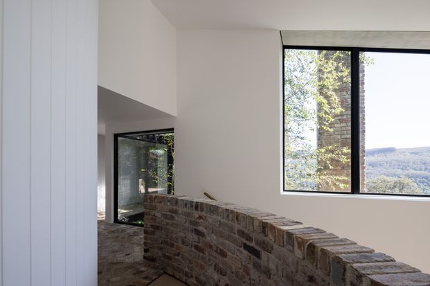 A bright, modern interior with a curved low-height exposed brick partition wall and dark-framed floor-to-ceiling glazing. The glazing opens views toward trees and a distant wooded landscape, while white walls and a clean-lined ceiling create a restrained architectural composition. A dark-framed glass doorway and brick flooring are visible in the adjoining area.