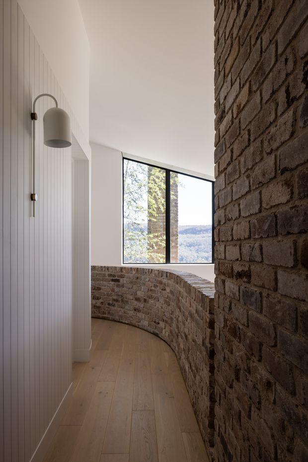 A narrow curved interior passage with a warm timber floor, exposed reclaimed-looking brick walls and a low curved brick balustrade. The left wall features vertical white panelling and a sculptural wall light, while a large black-framed window at the end provides views of trees and a hilly landscape. The space has a refined contemporary character with strong textural contrast between white panelling, brickwork and timber.